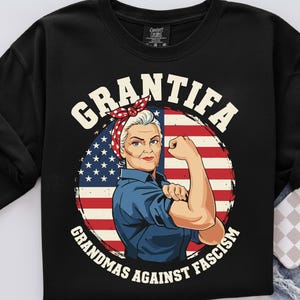 Peut inclure: Sweat-shirt noir avec un graphisme d'une femme montrant ses muscles devant un drapeau américain. Le texte "GRANTIFA GRANDMAS AGAINST FASCISM" est en blanc, en arc de cercle autour de l'image.