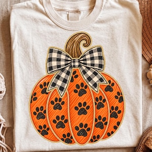 Puede incluir: Una camiseta color crema con un gráfico de una calabaza naranja grande y un lazo de cuadros blanco y negro. La calabaza tiene una superficie naranja texturizada con un patrón repetido de huellas de patas negras y un borde de purpurina dorada.