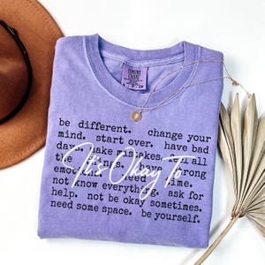 May include: A lavender short-sleeved t-shirt with a crew neckline. The shirt features black printed text that reads: be different. change your mind. start over. have bad days. make mistakes. feel all the things. have strong emotions. need time. not know everything. ask for help. not be okay sometimes. need some space. be yourself. Overlaid in white script is the text: It's Okay To.