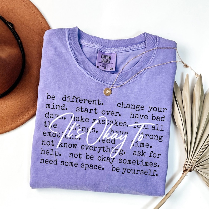 May include: A lavender short-sleeved t-shirt with a crew neckline. The shirt features black printed text that reads: be different. change your mind. start over. have bad days. make mistakes. feel all the things. have strong emotions. need time. not know everything. ask for help. not be okay sometimes. need some space. be yourself. Overlaid in white script is the text: It's Okay To.