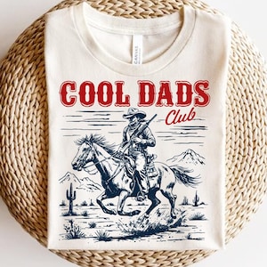 May include: A white t-shirt with a vintage-style graphic of a cowboy riding a horse. The graphic includes the text "Cool Dads Club" in red and blue.