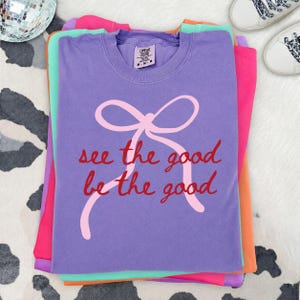 May include: A purple short-sleeve t-shirt featuring a light pink graphic of a tied ribbon bow. The text "see the good be the good" is printed in red cursive script across the center. The shirt has a Comfort Colors brand label at the neck.