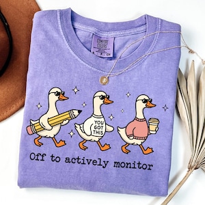 Può includere: Una maglietta lilla Comfort Colors con tre oche dei cartoni animati che indossano occhiali da sole. Un'oca tiene una matita, un'altra ha una maglietta con la scritta "You Got This" e la terza porta una tazza di caffè. Il testo "Off to actively monitor" è stampato in basso.