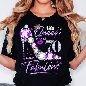 Op de afbeelding: Zwart T-shirt met een ontwerp van een hoge hak in paarse en zilveren strass. Het ontwerp bevat de tekst "This Queen Makes 70 Look Fabulous".