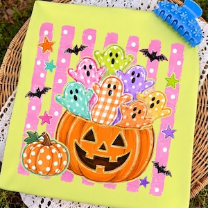 May include: A lime green t-shirt featuring a large orange jack-o'-lantern filled with colorful patterned ghosts. The background has pink vertical stripes with white polka dots, accented by small black bats and multi-colored stars.