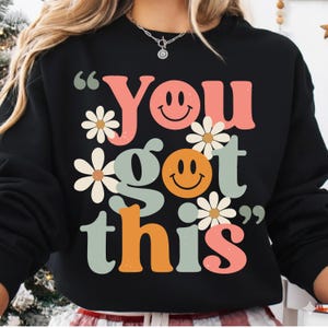 May include: Black sweatshirt with the phrase "You got this" in a retro font. The design includes smiley faces and daisy accents in shades of pink, orange, and green. A casual and motivational apparel item.