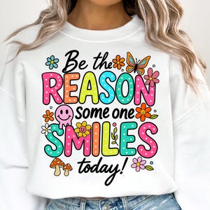 May include: White sweatshirt with a colorful graphic that says "Be the reason someone smiles today!" with flowers, a butterfly, and a smiley face.