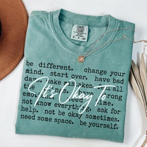 May include: A sage green short-sleeve t-shirt with a crew neckline and a Comfort Colors label. The shirt features black printed text that reads: be different. change your mind. start over. have bad days. make mistakes. feel all the things. have strong emotions. need time. not know everything. ask for help. not be okay sometimes. need some space. be yourself. Overlaid in white script is the text: It's Okay To.