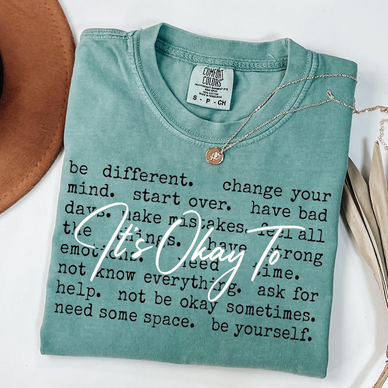 May include: A sage green short-sleeve t-shirt with a crew neckline and a Comfort Colors label. The shirt features black printed text that reads: be different. change your mind. start over. have bad days. make mistakes. feel all the things. have strong emotions. need time. not know everything. ask for help. not be okay sometimes. need some space. be yourself. Overlaid in white script is the text: It's Okay To.