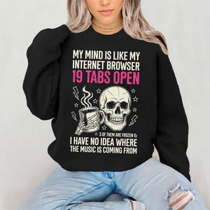 May include: Black sweatshirt with the text "MY MIND IS LIKE MY INTERNET BROWSER 19 TABS OPEN" in white and pink. The graphic includes a skull, a cup of coffee, and lightning bolts. The phrase "3 OF THEM ARE FROZEN & I HAVE NO IDEA WHERE THE MUSIC IS COMING FROM" is also printed on the sweatshirt.