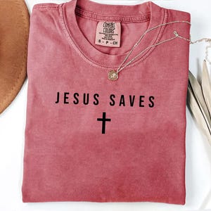 May include: A red t-shirt with the text "JESUS SAVES" and a cross underneath. The shirt is made of a soft, comfortable material.