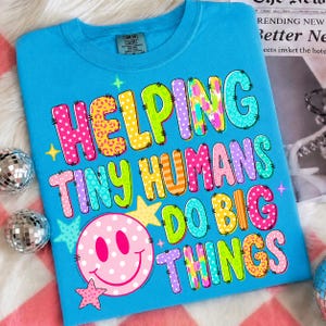 以下が含まれることがあります： 鮮やかなブルーのTシャツで、カラフルな柄入りのブロック体で「HELPING TINY HUMANS DO BIG THINGS」という文字がプリントされています。文字の下には、白い水玉模様の大きなピンクのスマイリーフェイスと小さなピンクの星が描かれています。