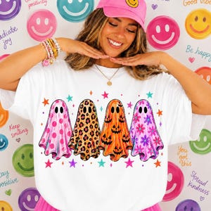 May include: A white t-shirt featuring a graphic print of four ghosts standing in a row. The first ghost has a pink polka dot pattern, the second has a leopard print, the third is orange with jack-o'-lantern faces, and the fourth has a floral pattern.