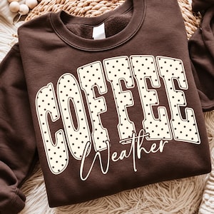 May include: A dark brown crewneck sweatshirt featuring the word COFFEE in large, cream-colored, polka-dotted block letters. Below the word COFFEE, the word Weather is printed in a white, cursive script font across the center of the chest.