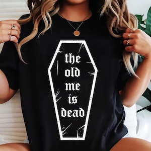 May include: Black t-shirt with a white coffin graphic and the words "the old me is dead" in a gothic font. The design is centered on the front. The shirt has short sleeves and a round neck.