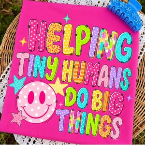 May include: A bright pink t-shirt featuring the colorful text HELPING TINY HUMANS DO BIG THINGS in various patterns like stars, leopard print, and polka dots. A large pink polka-dot smiley face and several small stars are printed on the shirt.