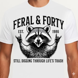 Op de afbeelding: Wit T-shirt met een zwart-wit wasbeer illustratie en de tekst "FERAL & FORTY EST. 1986 STILL DIGGING THROUGH LIFE'S TRASH". De wasbeer maakt het rock and roll handgebaar.