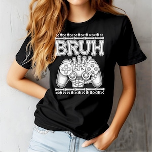 May include: Black t-shirt with a white graphic featuring the word "BRUH" in a bone-like font, a skeleton hand holding a video game controller, and skull and crossbones accents. The shirt is a crew neck.