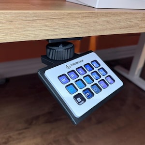 Może przedstawiać: Czarny Stream Deck z białym frontem i niebieskimi podświetlonymi przyciskami. Stream Deck jest zamontowany pod biurkiem za pomocą czarnego zacisku.