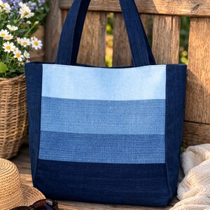 Könnte beinhalten: Eine Denim-Tragetasche mit Farbblock-Design in Blautönen. Die Tasche hat einen hellblauen oberen Bereich, einen mittelblauen mittleren Bereich und einen dunkelblauen unteren Bereich. Die Tasche hat zwei dunkelblaue Griffe.