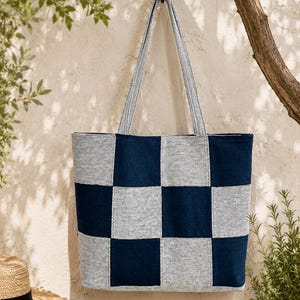 Könnte beinhalten: Eine große Tragetasche mit einem Schachbrettmuster in Marineblau und Hellgrau. Die Tasche hat lange Träger und hängt an einem Metallhaken. Ein Strohhut mit schwarzem Band ist im Hintergrund zu sehen.