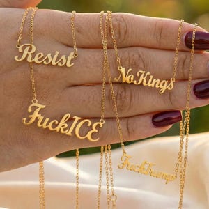 May include: Several gold necklaces are displayed, each with a different word: "Resist", "FuckICE", "No Kings", and "FuckTrump". The necklaces are held in a hand and have fine chains.