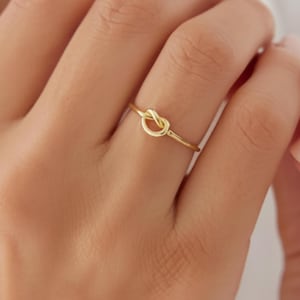 May include: A thin gold ring featuring a small, polished knot design at the centre. The band is smooth and circular, crafted from a reflective yellow gold metal. The knot element is integrated into the band, creating a continuous, minimalist loop.
