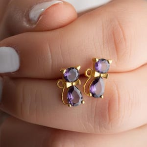 May include: A pair of gold-toned stud earrings shaped like cats, each featuring a round purple gemstone for the head and a pear-shaped purple gemstone for the body. The cat designs include small gold ears and a tail detail on the side of each earring.