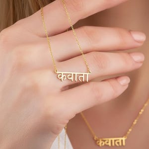 Könnte beinhalten: Eine goldfarbene Metallhalskette mit einem horizontalen Stabanhänger, auf dem der Text "कवाता" in Devanagari-Schrift eingraviert ist. Der Anhänger ist an einer zarten goldfarbenen Kette befestigt, die am Hals anliegt und von einer Hand gehalten wird.