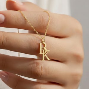 Peut inclure: Un délicat collier doré avec les initiales "BK". Le pendentif est suspendu à une fine chaîne, tenue par une main aux ongles manucurés. Le collier est un bijou simple mais élégant.
