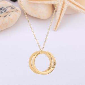 Könnte beinhalten: Eine goldfarbene Halskette mit zwei ineinandergreifenden Kreisen. Auf den Kreisen sind die Namen "Maicoo" und "Olivia" eingraviert. Die Kette wird auf einer weißen Oberfläche mit Muscheln und einem Seestern präsentiert.