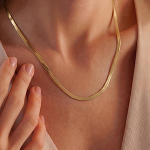 May include: A gold-toned herringbone chain necklace featuring a flat, flexible link design that rests against the collarbone. The necklace has a polished, reflective surface and a smooth, continuous texture that catches the light along its length.