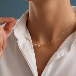 Könnte beinhalten: Eine zarte goldene Halskette mit einer Reihe kleiner, runder Perlen und einer dünnen Kette. Die Halskette wird vor einem weißen Hemd mit Kragen getragen, was einen Kontrast zum Gold bildet. Die Halskette wird von einer Hand gehalten.