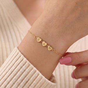 Può includere: Un bracciale a catena color oro con tre piccoli ciondoli a forma di cuore disposti in fila. Ogni cuore è inciso con una lettera maiuscola, nello specifico E, A e M, centrata sulla superficie dei cuori in metallo dorato.