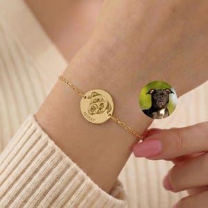 May include: A gold-toned chain bracelet featuring a circular pendant engraved with a line-art illustration of a dog's face and the name ASHLEY. A small circular inset shows a photograph of a black dog with brown markings.