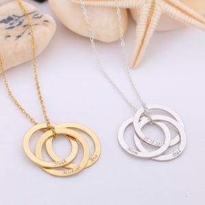 Może przedstawiać: Dwa naszyjniki z zazębiającymi się okręgami. Jeden jest w kolorze złotym, wygrawerowany imionami "Noah", "Michael" i "Ava". Drugi jest w kolorze srebrnym, wygrawerowany napisami "Mom", "Dad" i datą. Naszyjniki są prezentowane na białej powierzchni z muszlami.