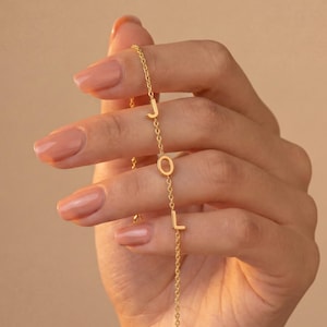Pode incluir: Uma pulseira de corrente em tom dourado com três pingentes de letras maiúsculas orientados verticalmente que formam a palavra JOL. Os delicados elos da corrente de metal são intercalados com as letras J, O e L, criando um design linear.