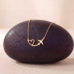 May include: A gold-colored necklace featuring a heart and airplane pendant. The pendant is connected to a delicate chain. The necklace is displayed on a dark, textured stone against a soft, neutral background.
