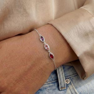 Könnte beinhalten: Ein zartes Silberkettenarmband mit drei in einer Reihe angeordneten, marquise-geschliffenen Edelsteinen. Die Steine sind in einem Farbverlauf angeordnet, beginnend mit einem hellvioletten Amethyst, gefolgt von einem zartrosa Stein und einem tiefroten Edelstein.
