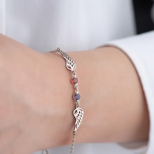 Puede incluir: Una pulsera de cadena en tono plateado con dos dijes simétricos en forma de ala y dos piedras preciosas redondas engastadas en rojo y morado. La delicada cadena de metal conecta las alas y las piedras en una disposición lineal y elegante.