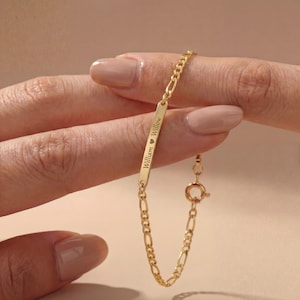 May include: A gold bracelet featuring a Figaro chain and a rectangular bar. The bar is engraved with the names "William" and "Willow" separated by a heart symbol. The bracelet is fastened with a circular clasp.