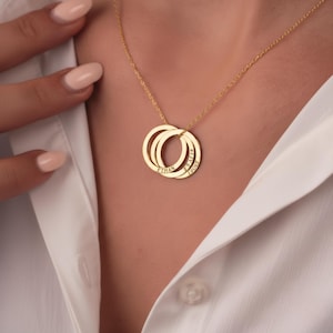 May include: A gold-toned necklace featuring three interlocking circular pendants. Each circular pendant is engraved with a name, specifically Ethan, Sofia, and Lucas, suspended from a delicate gold-toned chain that rests against a white collared shirt.