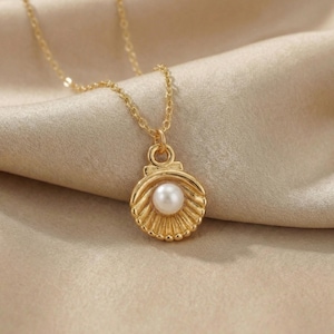 Könnte beinhalten: Eine goldfarbene Halskette mit einem Muschel-Anhänger. Der Anhänger hat eine weiße Perle in der Mitte, umgeben von einem detaillierten, strukturierten Muscheldesign. Die Halskette ist filigran und ergänzt den eleganten Stil des Anhängers.