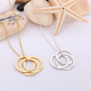 Op de afbeelding: Twee kettingen, een goudkleurige en een zilverkleurige, elk met drie in elkaar grijpende ronde hangers. De gouden ketting is gegraveerd met de namen Noah, Michael en Ava. De zilveren ketting is gegraveerd met Mom, Dad en 25.12.2020.