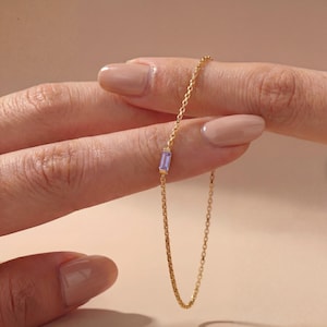 May include: A delicate gold chain bracelet featuring a single, small, rectangular, light purple baguette-cut gemstone set in a gold frame. The chain consists of thin, interlocking circular links, creating a refined and simple jewelry piece.