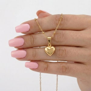 May include: A gold-toned necklace featuring a small, smooth, puffed heart-shaped pendant suspended from a delicate chain with evenly spaced small gold beads. The metal has a polished, reflective finish, and the heart hangs centrally from the chain.