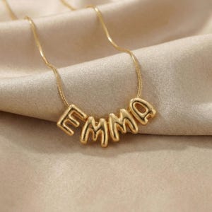 Könnte beinhalten: Eine goldfarbene Halskette mit dem Namen "EMMA" in Blockbuchstaben. Die Halskette wird auf einem beigen Satinstoff präsentiert, wobei die Kette und die Buchstaben aus Metall zu bestehen scheinen.