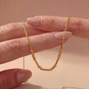 May include: A gold-toned necklace featuring a delicate chain interspersed with a repeating pattern of small round beads and thin cylindrical bars. The design creates a linear sequence of metallic elements that drape in a gentle curve.