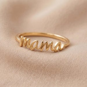 Könnte beinhalten: Ein goldfarbener Ring mit dem Wort mama in durchgehender Schreibschrift. Das Metall hat eine glatte, polierte Oberfläche und die Buchstaben sind direkt in das Ringband eingearbeitet, was ein einheitliches Schmuckstück ergibt.