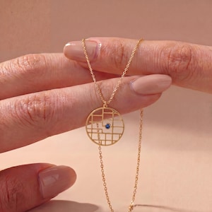 May include: A gold-toned necklace featuring a circular pendant with an open grid pattern and a single small blue gemstone set in the center. The pendant hangs from a delicate gold-toned chain, creating a geometric design with a central blue accent.